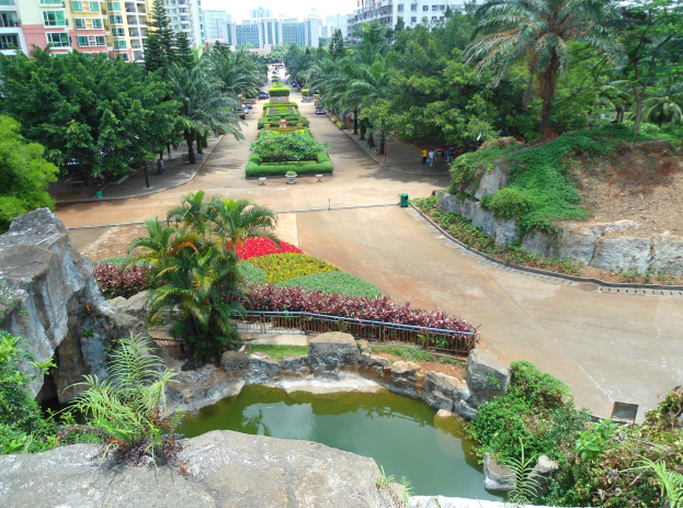 Luftaufnahme eines Parks mit einem Teich, umgeben von Felsen, Pflanzen, Bäumen und Blumen, Gebäuden im Hintergrund und Menschen, die darunter herumlaufen.