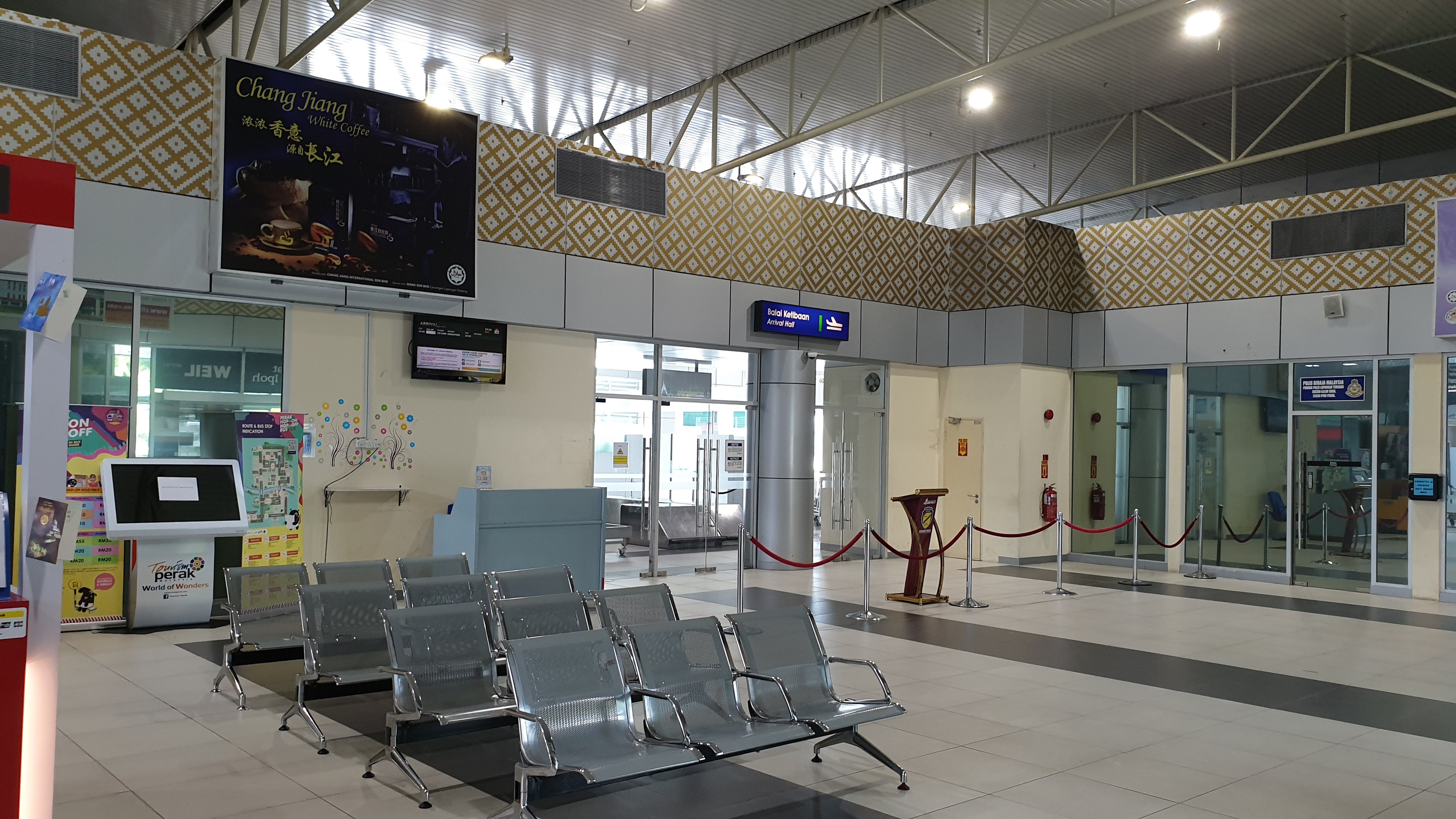 Leerer Flughafen-Wartebereich mit Stühlen auf dem Boden, Absperrpoller mit Seilen, Informationsanzeigen, wandmontierte Bildschirme, Deckenleuchten und Glastüren.