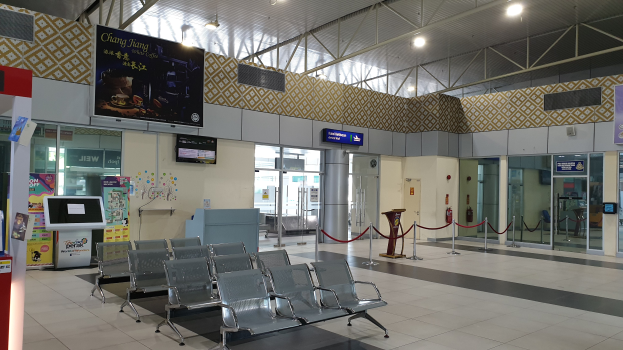 Leerer Flughafen-Wartebereich mit Stühlen auf dem Boden, Absperrpoller mit Seilen, Informationsanzeigen, wandmontierte Bildschirme, Deckenleuchten und Glastüren.