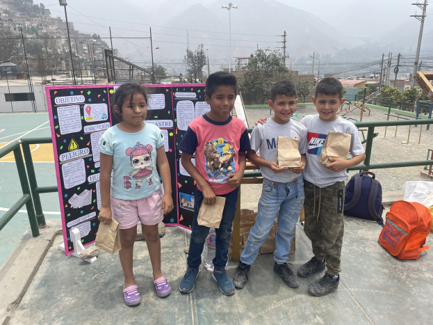 Gruppe von Kindern auf einem Basketballfeld mit Papiertüten in den Händen, Tüten und Gegenstände rechts und eine Tafel mit Text links, vor einem Hintergrund aus Zaun, Pfosten, Laternen, Strommasten, Bäumen, Gebäuden, Hügeln und Himmel.