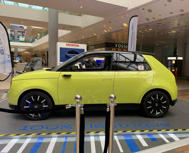 Ein gelbes Wasserstoff-elektrisches Auto steht vor einem Gebäude mit Absperrpollern, Bannern, Menschen, Schildern, einer Topfpflanze, Deckenleuchten und Ventilatoren.