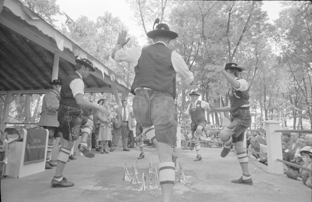Eine Gruppe Männer in traditioneller bayrischer Tracht tanzt auf einer Bühne vor einem gemischten Publikum, mit Bäumen und einem Schuppen im Hintergrund, dargestellt in Schwarz-Weiß.
