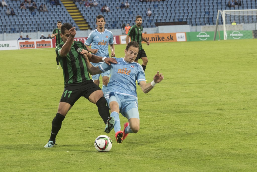 Menschen spielen ein Ballspiel auf Rasen mit Netz und Tribünen, Zuschauer sind im Hintergrund eines Stadions mit Stühlen sichtbar.