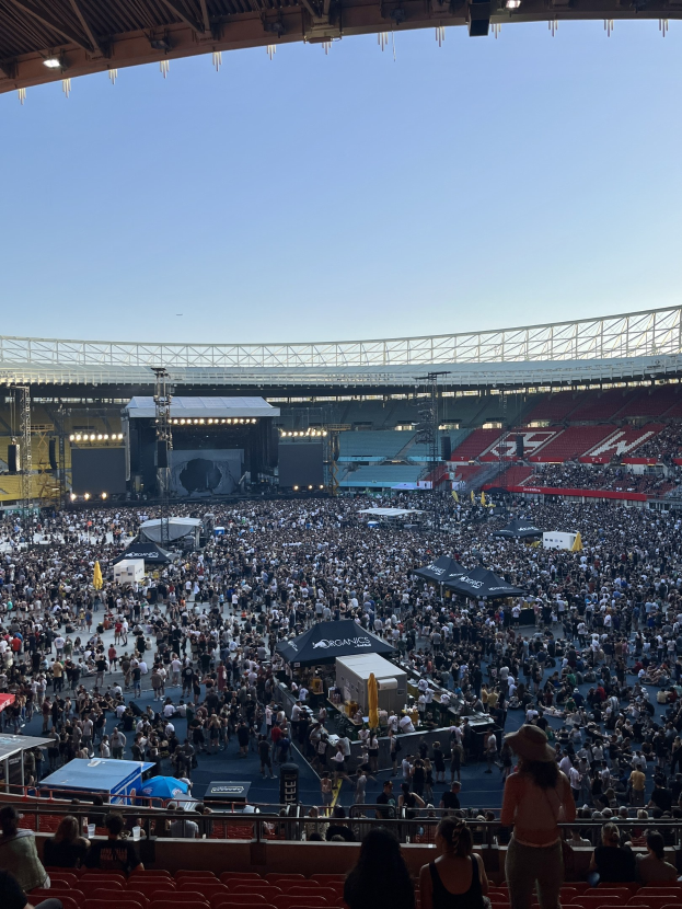 Große Menschenmenge auf Sitzplätzen und Stehplätzen in einem Stadion mit Zelten, Geländern und Überdachung unter einem Himmelhintergrund, was auf ein Konzert in der O2 Arena in London hinweist.