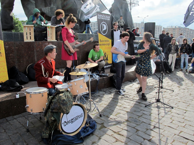 Eine Gruppe von Menschen spielt Musikinstrumente auf einem Gehweg mit verstreuter Ausrüstung und Bannern im Hintergrund.