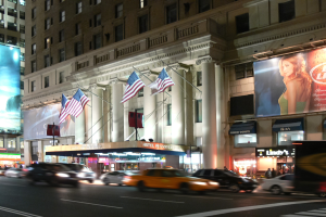 Lebendige Stadtstraße bei Nacht mit fahrenden Fahrzeugen, ein mehrstöckiges Gebäude mit beleuchteten Fenstern, Säulen und Fahnenmasten, beleuchtete Informationsschilder mit einem Frauenbild und Straßenbeleuchtung.