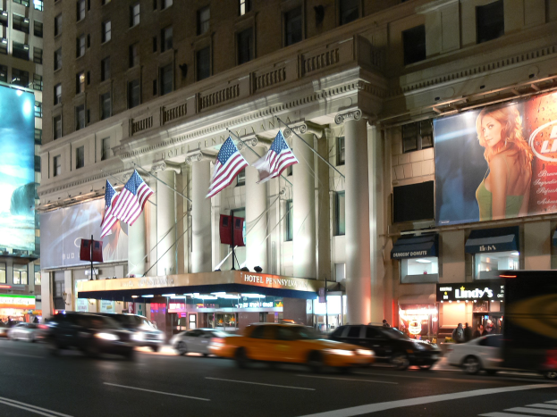 Lebendige Stadtstraße bei Nacht mit fahrenden Fahrzeugen, ein mehrstöckiges Gebäude mit beleuchteten Fenstern, Säulen und Fahnenmasten, beleuchtete Informationsschilder mit einem Frauenbild und Straßenbeleuchtung.