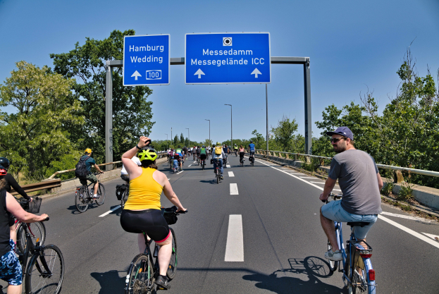 Gruppe von Radfahrern in Helmen auf einer Straße mit einer Begrenzung auf der linken Seite und Bäumen auf der rechten Seite, Laternen im Hintergrund, unter einem klaren blauen Himmel, mit einem Schild, das eine Radtour in Hamburg anzeigt.