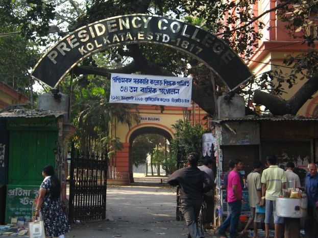Der Eingang zum Presidency College in Kolkata, Indien, mit einem Torbogen mit einem "Presidency College"-Schild, einem Tor, Menschen, die rechts gehen, einem Gebäude, Bäumen und einem klaren blauen Himmel im Hintergrund.