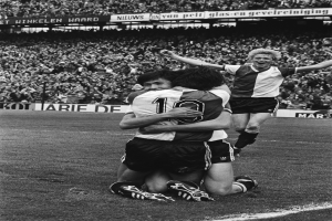 Drei Fußballspieler feiern ein Tor auf dem Boden vor einer jubelnden Menge in einem schwarz-weißen Stadion.