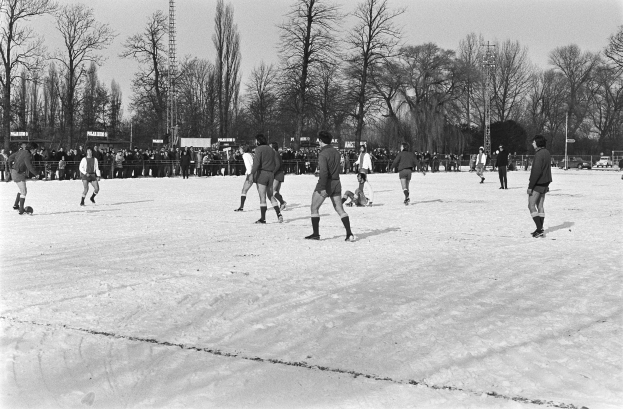Schwarzes und weißes Bild von Menschen, die auf einem schneebedeckten Feld mit Bäumen, Pfählen und einem klaren Himmel Fußball spielen.