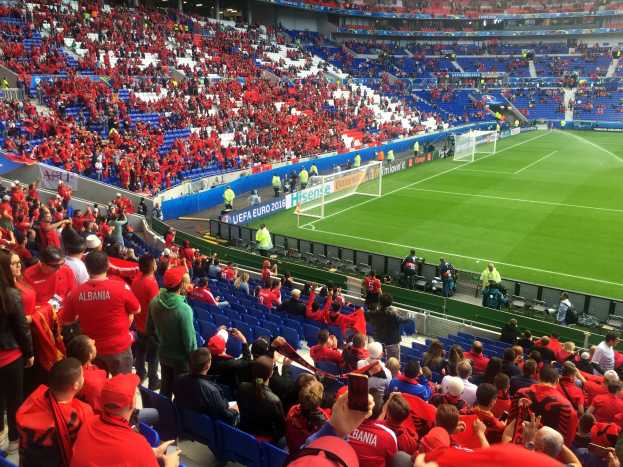 Eine große Menschenmenge in einem Stadion schaut bei einem Fußballspiel zu, mit Sitzplätzen und Stehplätzen, Fahnen, einem Torpfosten und einer Decke mit Leuchten.