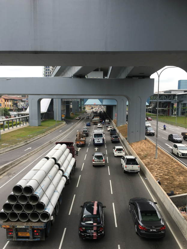 Eine vielbefahrene Autobahn mit mehreren Fahrzeugen, eine Brücke darüber, Straßenlaternen, Gras, Gebäude, Bäume und ein Himmel-Hintergrund.